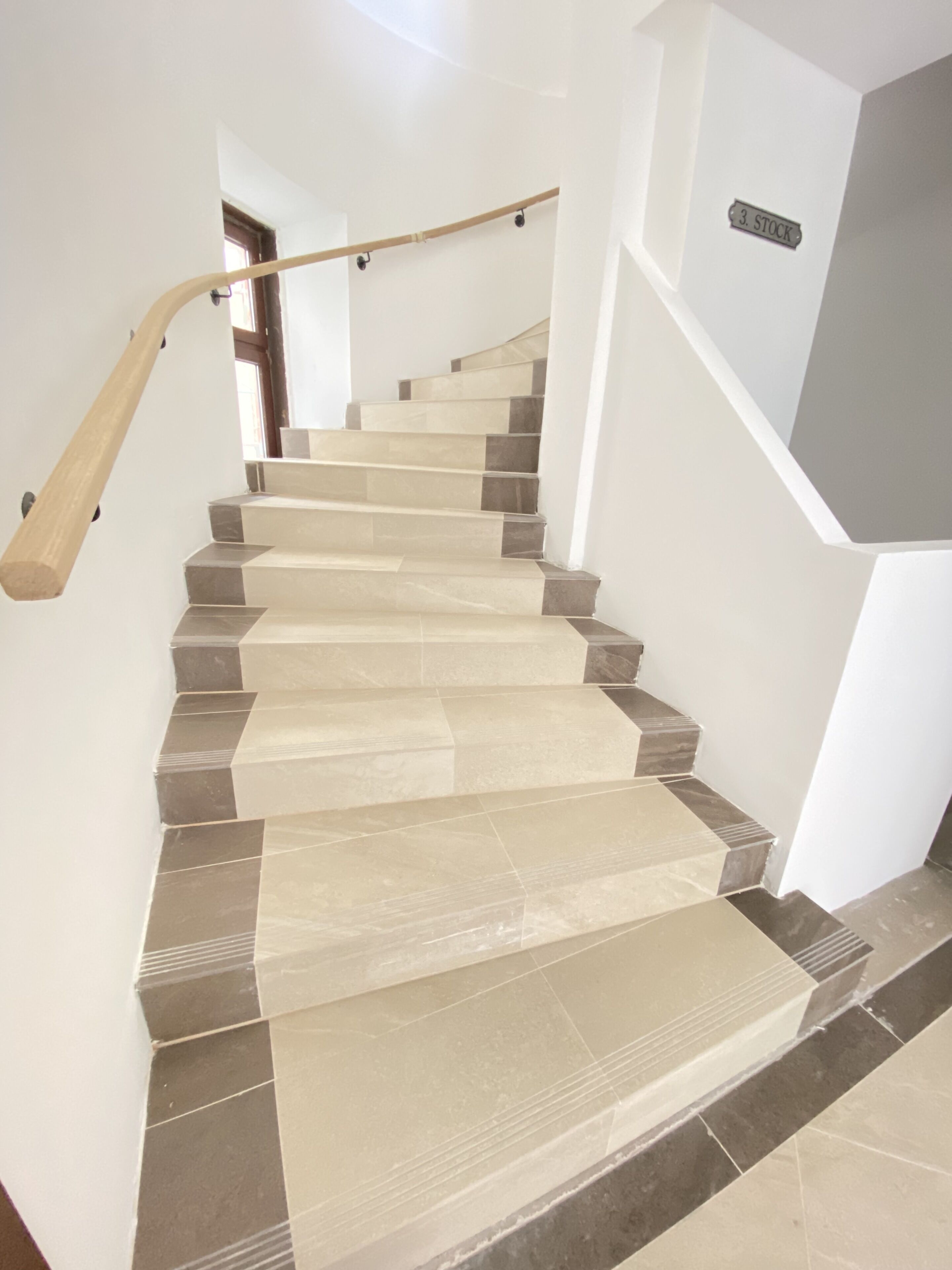 Una escalera con baldosas color beige y gris, un pasamanos de madera y un letrero que dice '3. STOCK'.