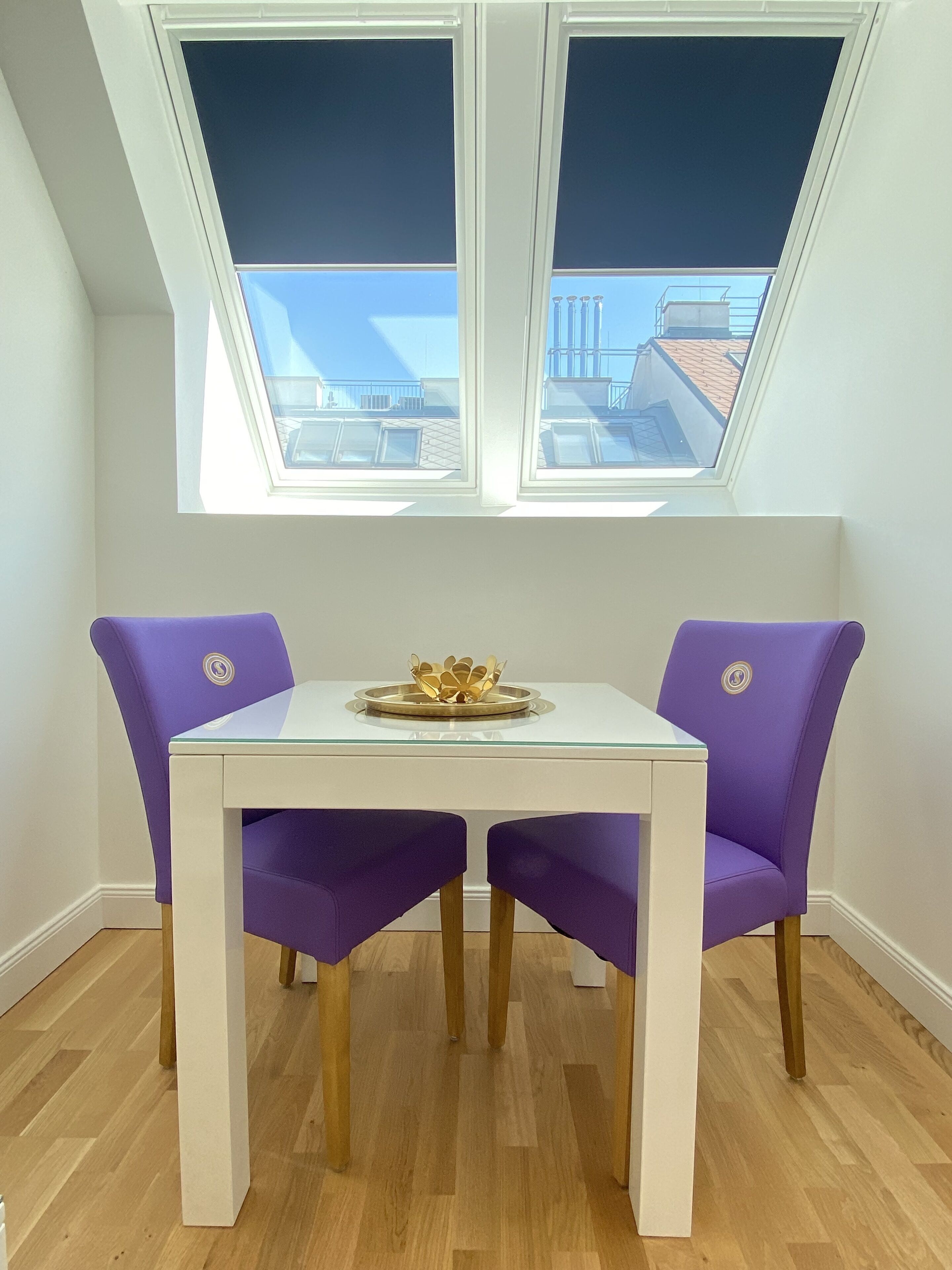 A dining area with a white table and purple chairs, a gold decorative bowl on the table, and a view of buildings through skylights.