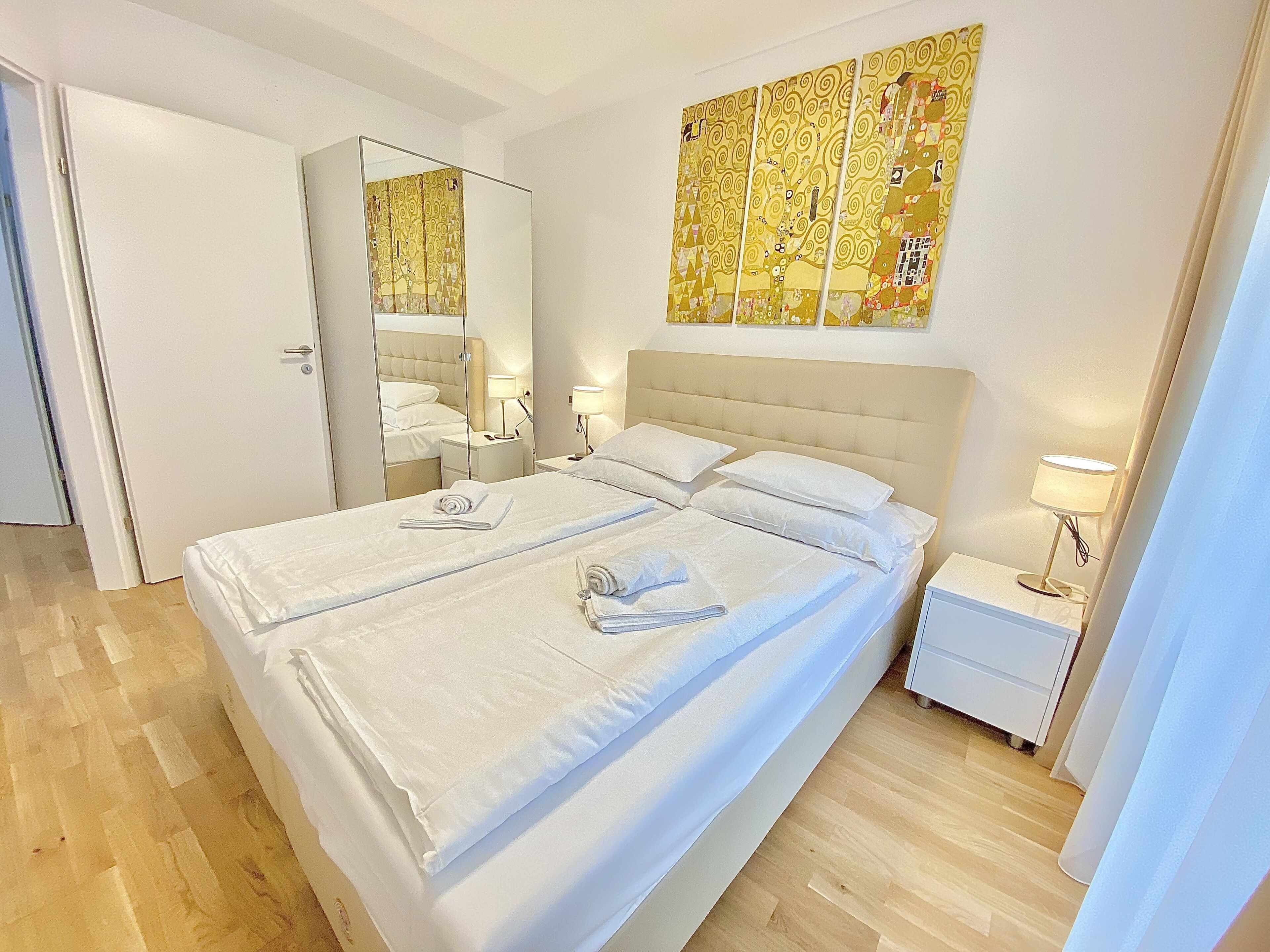A hotel room with a large bed, a bedside table, a nightstand, a mirror, and a wall art piece.