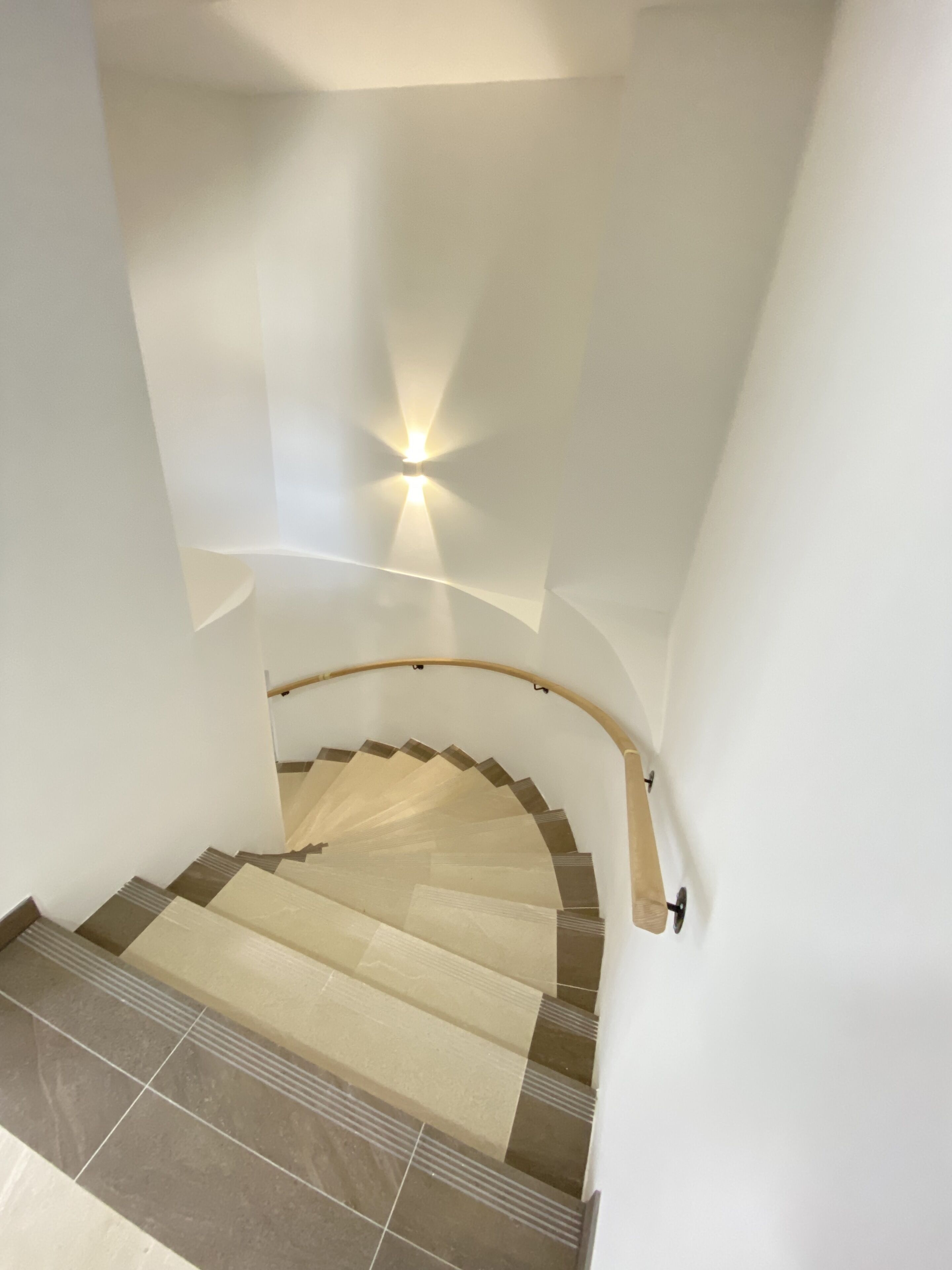 A staircase with a wooden handrail and beige steps, leading up to a landing with a wall-mounted light fixture.