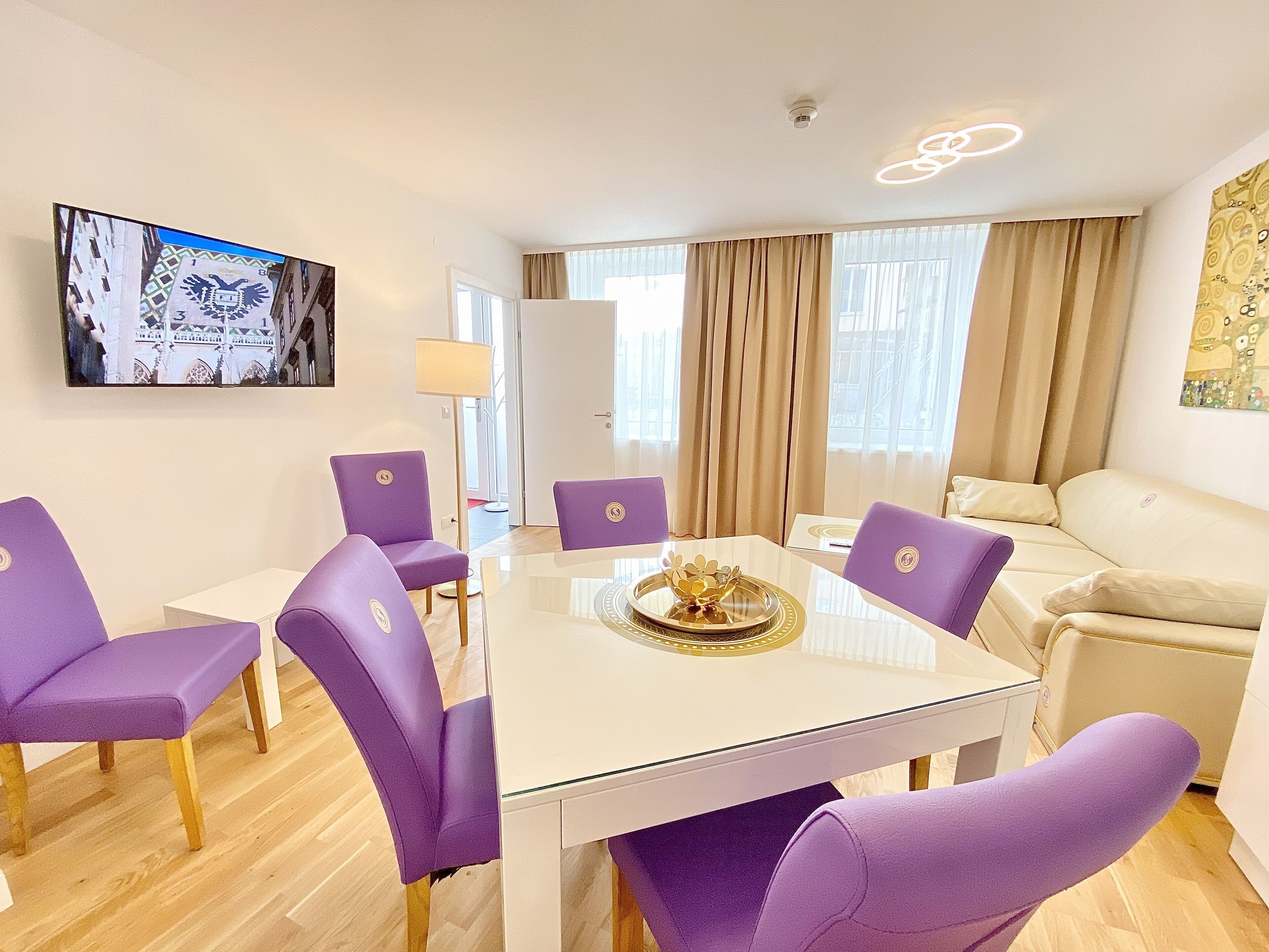 A modern hotel room with a dining area featuring a glass table and purple chairs, a beige sofa, and a large window with curtains.