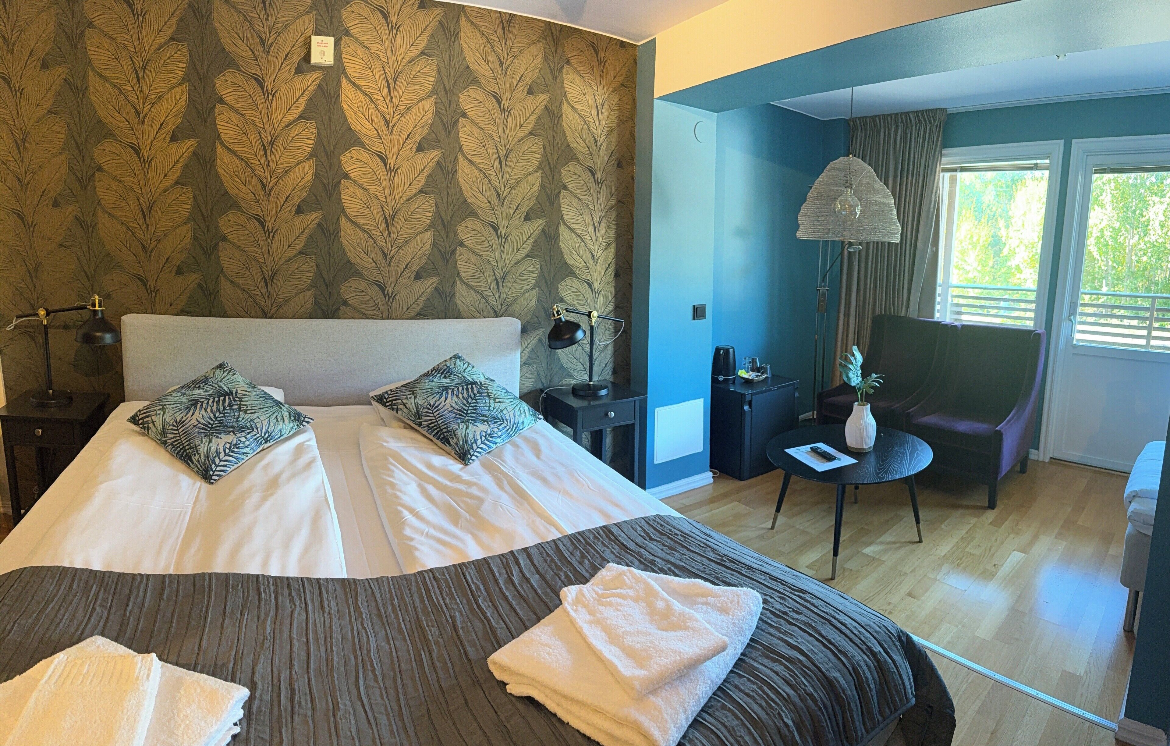 Habitación de hotel con una cama grande, dos almohadas, una mesita de noche con lámpara, una silla, una pequeña mesa con un florero y vistas a la vegetación por la ventana.