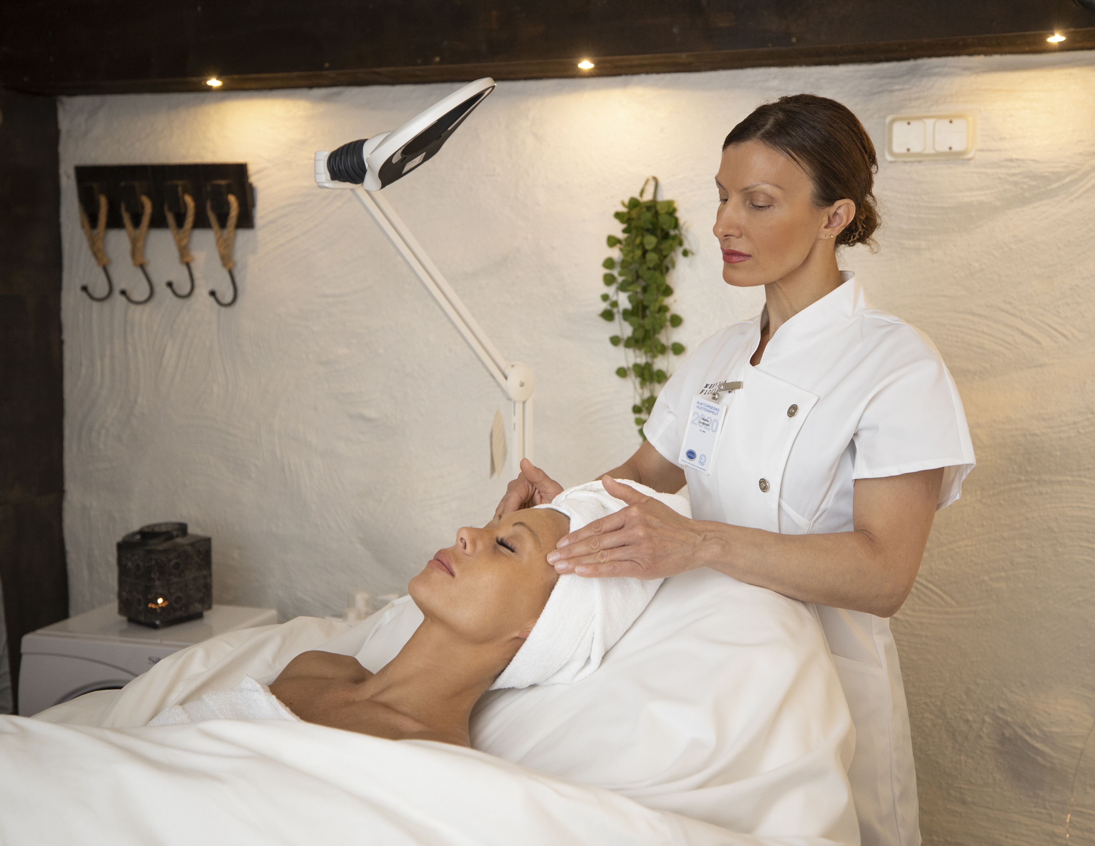 A person lying on a bed receiving a facial treatment in a room with a white wall, a plant, and a wall-mounted lamp.