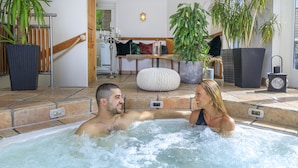 A couple in a hot tub with a stone floor, surrounded by potted plants and a wooden bench.