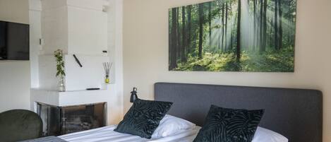 Una habitación de hotel moderna con una cama grande, dos almohadas oscuras, un cabecero blanco, un cuadro de un bosque, una mesita y una silla.