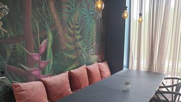 Uma área de refeições com mesa e assentos acolchoados, um mural com motivos florestais e de animais, e uma janela com cortinas leves.