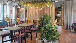 A modern restaurant interior with wooden tables, yellow chairs, hanging light bulbs, and potted plants.