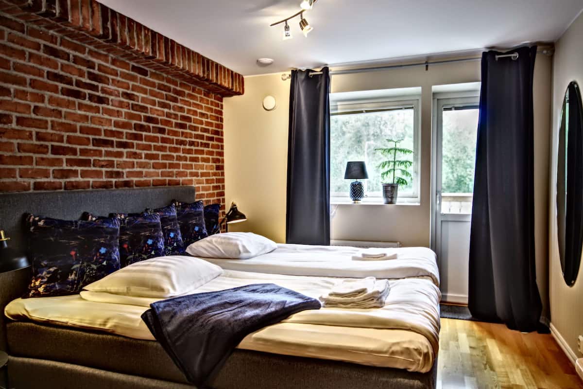 Une chambre d’hôtel avec un mur en briques, deux lits, une fenêtre avec des rideaux, une plante et une lampe.