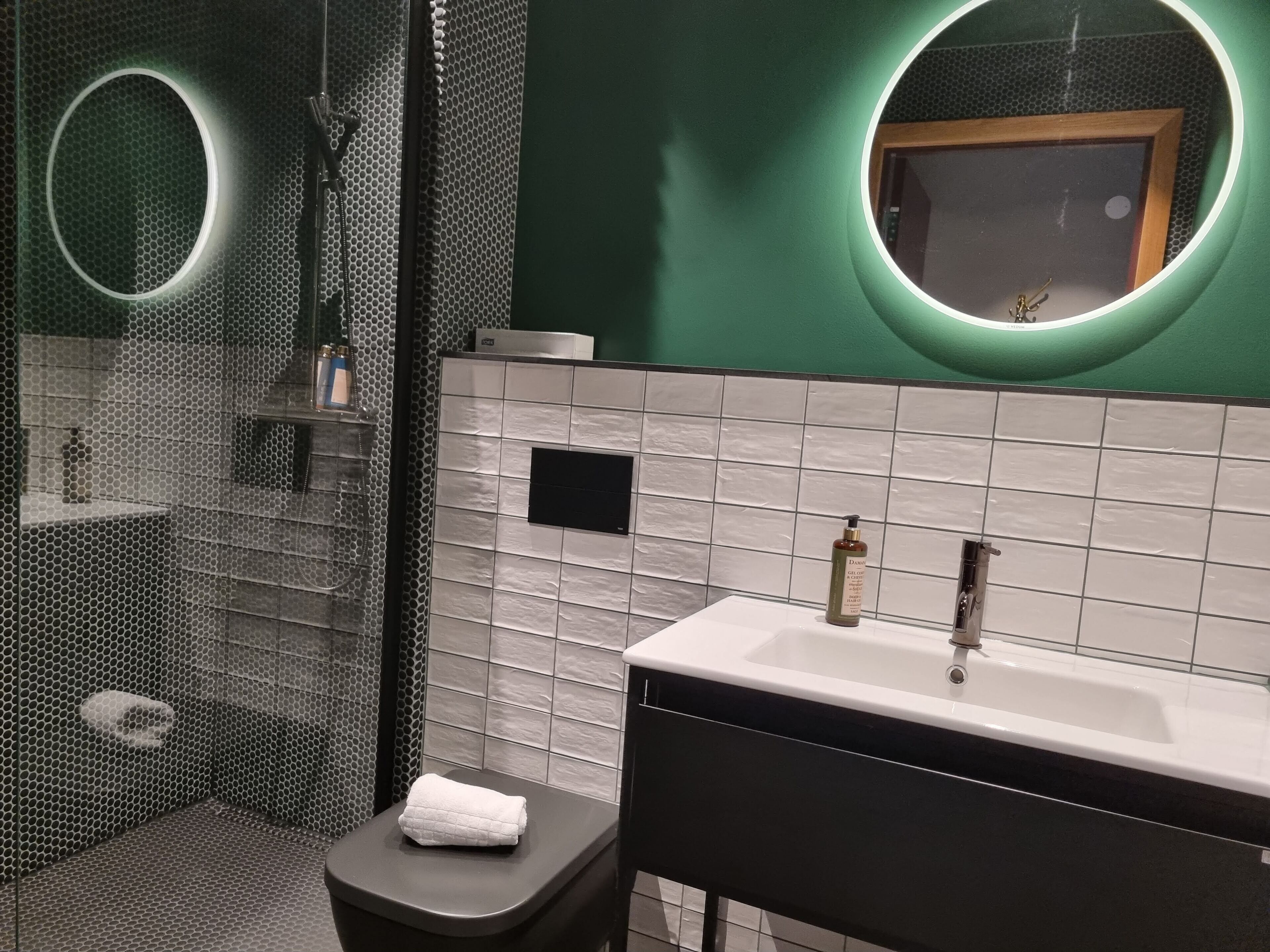 Un baño moderno con cabina de ducha de vidrio, un lavamanos blanco sobre encimera negra y un espejo con marco circular.