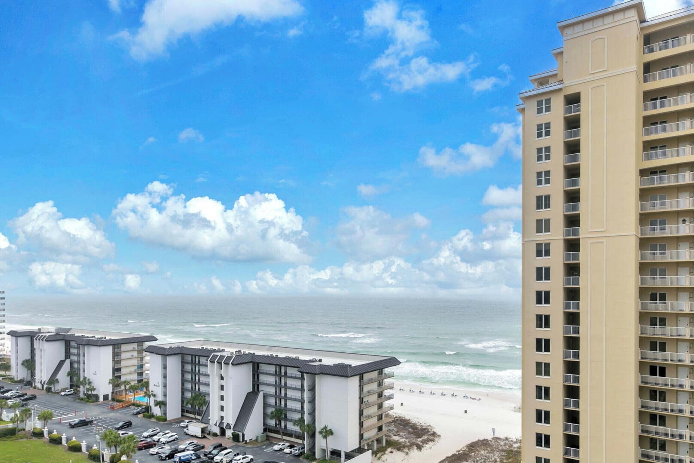 A coastal hotel with multiple floors, a parking lot, and a beach view.