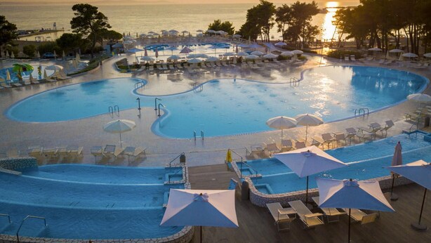 A large, multi-level swimming pool with sun loungers and umbrellas, surrounded by palm trees and a view of the ocean at sunset.