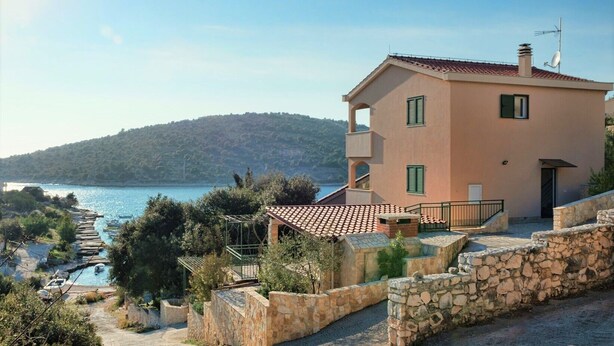 Un edificio de dos plantas con un muro de contención de piedra, una piscina pequeña y vistas a un lago y colinas.