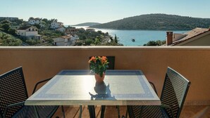 Un balcón con mesa y sillas, una planta en maceta y vista a una zona costera con edificios y colinas.