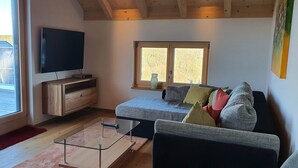 A modern living room with a large TV, wooden furniture, a glass coffee table, and a window with a view of a wooden deck. - Adlernest (Feldberg)