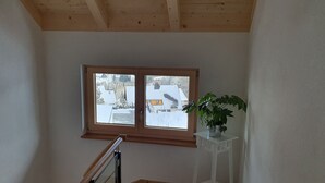 A room with a wooden ceiling, a window with a view of a snowy landscape, and a potted plant on a white stand. - Adlernest (Feldberg)