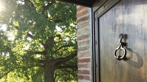Eine Holztür mit Metallbeschlägen, eine Ziegelwand und ein Blick auf einen Baum und eine Schotterfläche.
