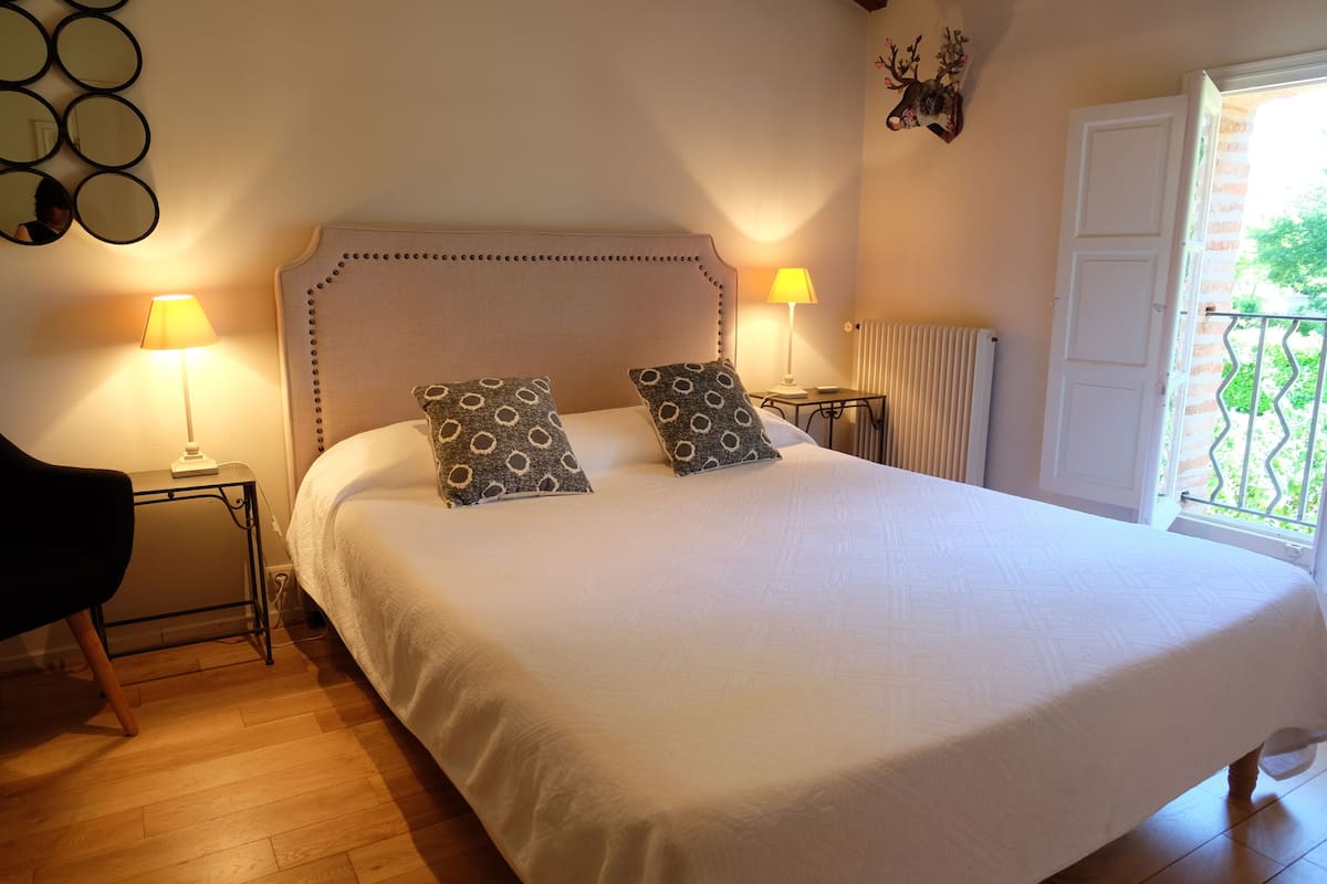 Una cama bien hecha con almohadones estampados, dos lámparas de noche, una mesita y un adorno en la pared.