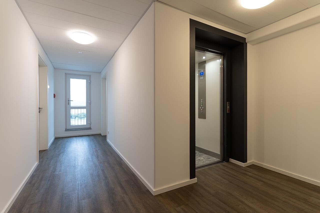 A hallway with a wooden floor, a door leading to an elevator, and a window at the end.
