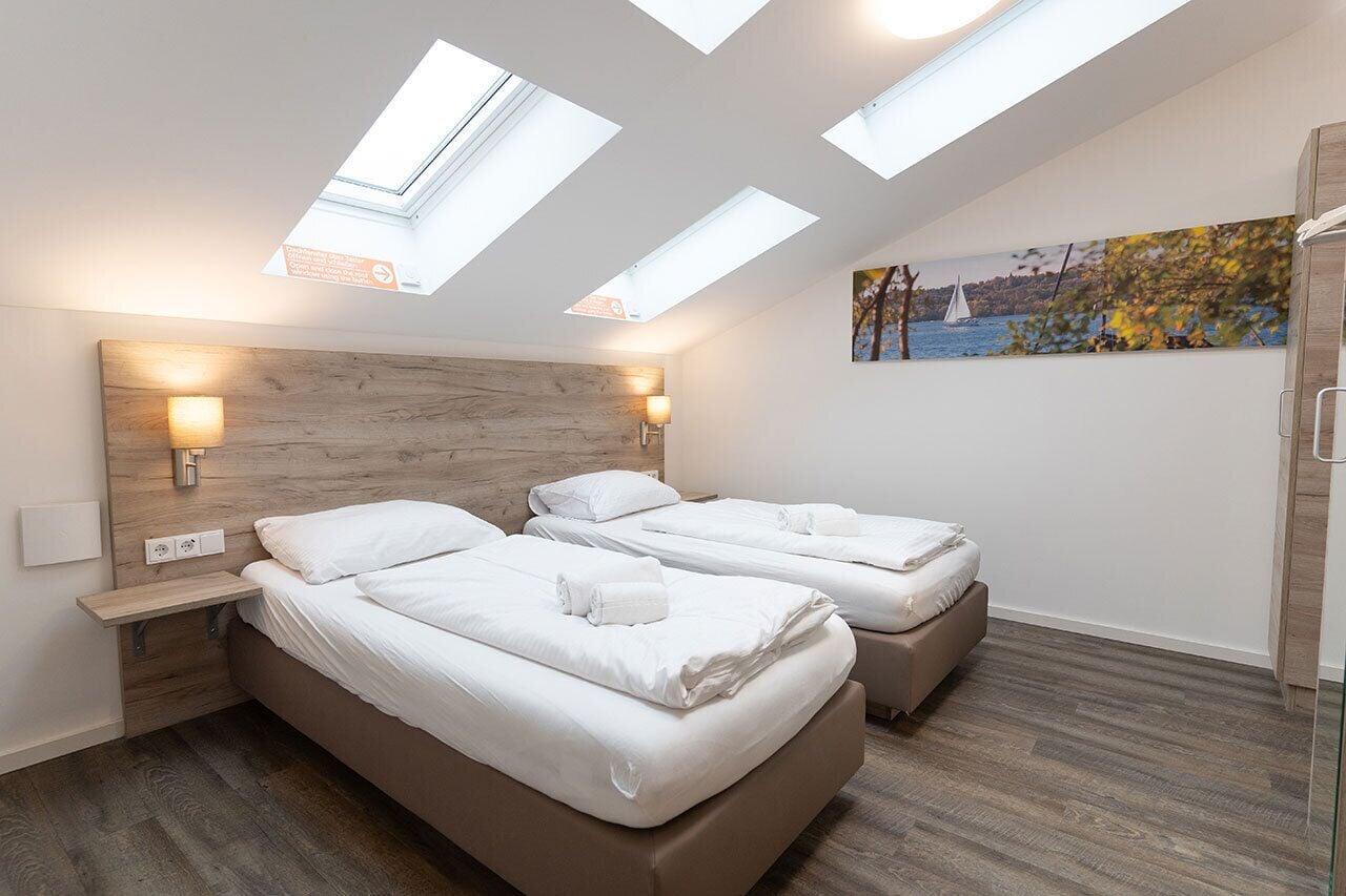 Une chambre moderne avec deux lits, des fenêtres de toit, une tête de lit en bois et un tableau représentant un voilier.