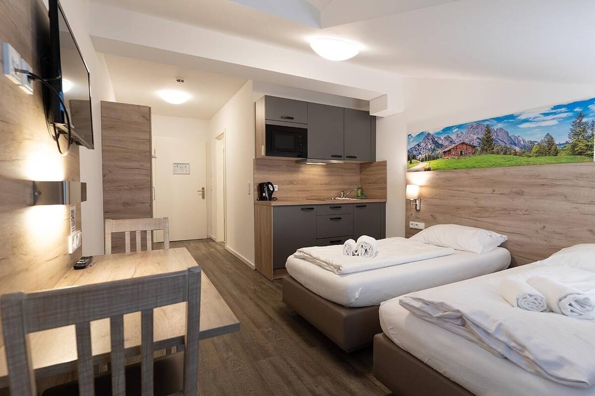 A hotel room with a wooden floor, a desk with a chair, a bed with white linens, a microwave, and a painting of mountains on the wall.