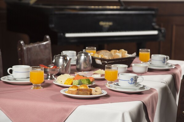 A breakfast setting with a piano in the background, a table with plates of food, cups, and a pitcher of orange juice. - Vitoria Palace Hotel (Lavras)