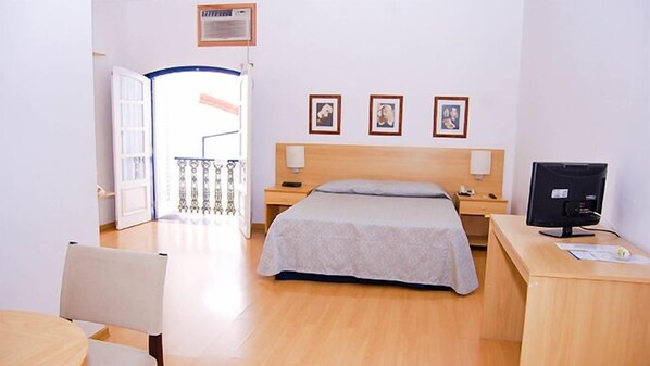 A hotel room with a bed, bedside tables, a desk with a computer, and a chair. There are framed pictures on the wall and a balcony door. - Vitoria Palace Hotel (Lavras)