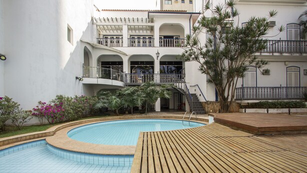 A swimming pool with a wooden deck, surrounded by a white building with balconies and a garden with pink flowers. - Vitoria Palace Hotel (Lavras)