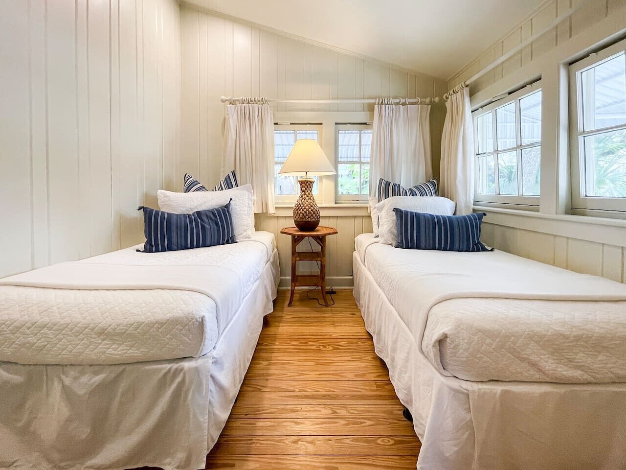 A room with two beds, a small table with a lamp, and large windows with curtains.