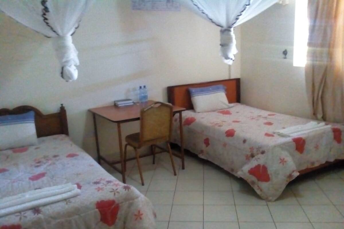 A room with two single beds, a wooden desk, a chair, and a small table with a water bottle.