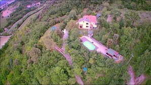 An aerial view of a house surrounded by dense greenery, with a swimming pool and a tennis court visible.