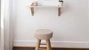 Ein Zimmer mit weißer Wand, einem Holzschemel, einem runden Spiegel und einem kleinen Regal mit Pflanze und Kerze.