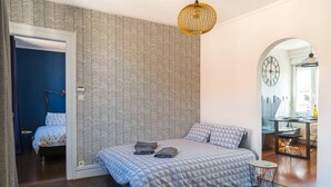 Ein Schlafzimmer mit strukturierter Wandtapete, einem Bett mit geometrischem Muster, einer Nachttischlampe und einem kleinen, runden Tisch mit Stuhl.