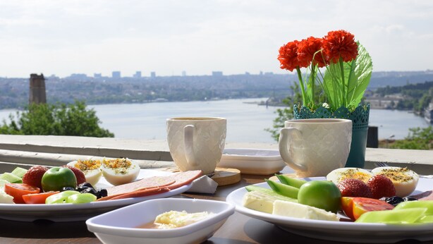 A breakfast setting with a scenic view of a river and city skyline, including a table with plates of food, cups, and a vase with flowers. - Galata Dream Hotel (Istanbul)