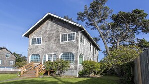 A two-story house with a wooden staircase leading to the front door, surrounded by a well-maintained lawn and a large tree. - Enjoy This 4 Bedroom w/ a Beautiful Yard, Ocean Views and Easy Beach Access! (Seaside)