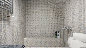 Una doccia piastrellata con soffitto curvo, un miscelatore a doccia manuale e uno scaffale con articoli da toilette.