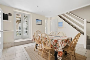 Comedor con una mesa lista para cuatro personas, rodeada de sillas de madera, un mantel estampado y vista a una escalera.