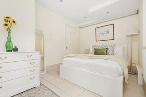 A bedroom with a white bed, a dresser with drawers, a vase with flowers, a framed picture on the wall, and a lamp.