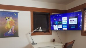 Una stanza con una scrivania, una sedia, una TV a muro che mostra diversi servizi di streaming, una finestra con una tenda e un quadro appeso al muro.