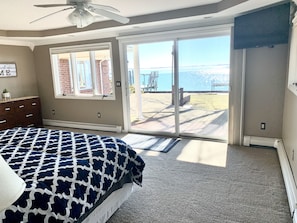 A bedroom with a bed, a dresser, a ceiling fan, and a sliding glass door leading to an outdoor deck with a view of the ocean. - Beautiful Lakefront Home On Lake St Clair! (Ira)
