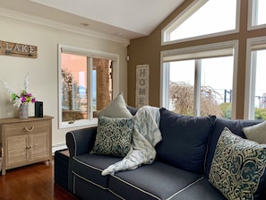 A cozy living room with a dark blue sofa, patterned cushions, and a decorative vase with flowers. A wooden cabinet with a drawer is visible, and a large window offers a view of the outdoors. - Beautiful Lakefront Home On Lake St Clair! (Ira)