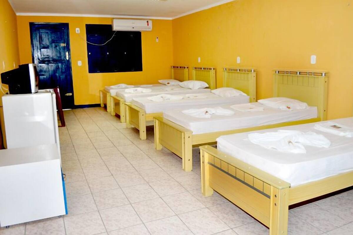 A hotel room with four single beds, a small kitchenette, and a door leading to a dark room.