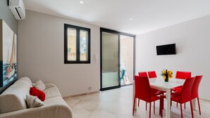 Una sala de estar moderna con un sofá, una mesa de comedor con sillas rojas, un televisor de pantalla plana y un ventanal corredero que da a un balcón.