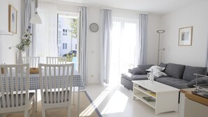 A bright living room with a white sofa, a dining table with chairs, and a large window with curtains.