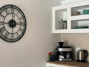 A kitchen with a wall-mounted clock, a coffee maker, a kettle, and shelves with dishes.