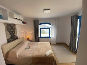 A bedroom with a bed, a window with curtains, an air conditioning unit, and a wall-mounted shelf with items.