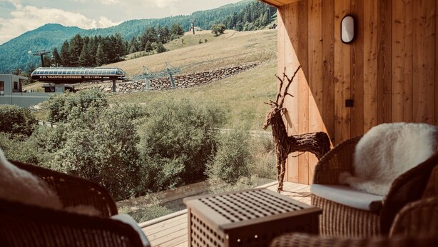 A wooden deck with wicker chairs, a deer's head sculpture, and a view of a mountain and ski lift. - Chalet 'Chalet Ortles' with Lake View, Private Terrace, and Wi-Fi (Sankt Valentin auf der Haide)