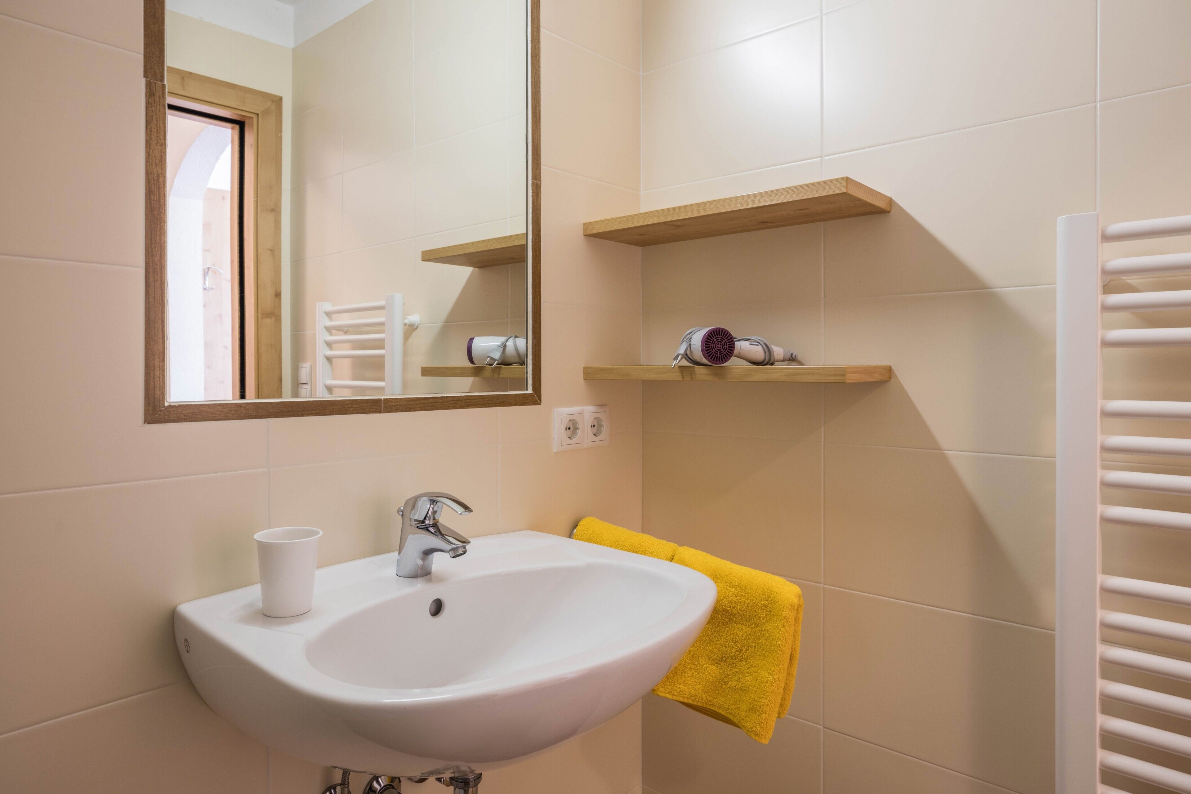 Une salle de bain avec un lavabo blanc, un miroir, un porte-serviettes et une étagère sur laquelle se trouve un sèche-cheveux.