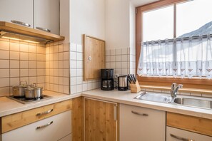 Uma cozinha com fogão, cafeteira e utensílios sobre a bancada, armários de madeira e uma janela com cortina.