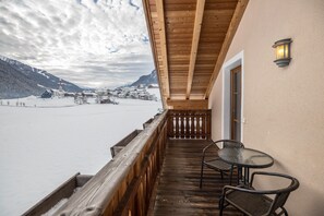 Un balcón con baranda de madera, una mesa y dos sillas, con vista a un paisaje nevado y montañas al fondo.