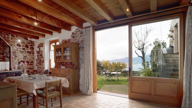 Un coin repas avec une table dressée pour deux personnes, des poutres en bois au plafond, un mur en pierre et une vue sur un jardin, visible à travers une porte vitrée.